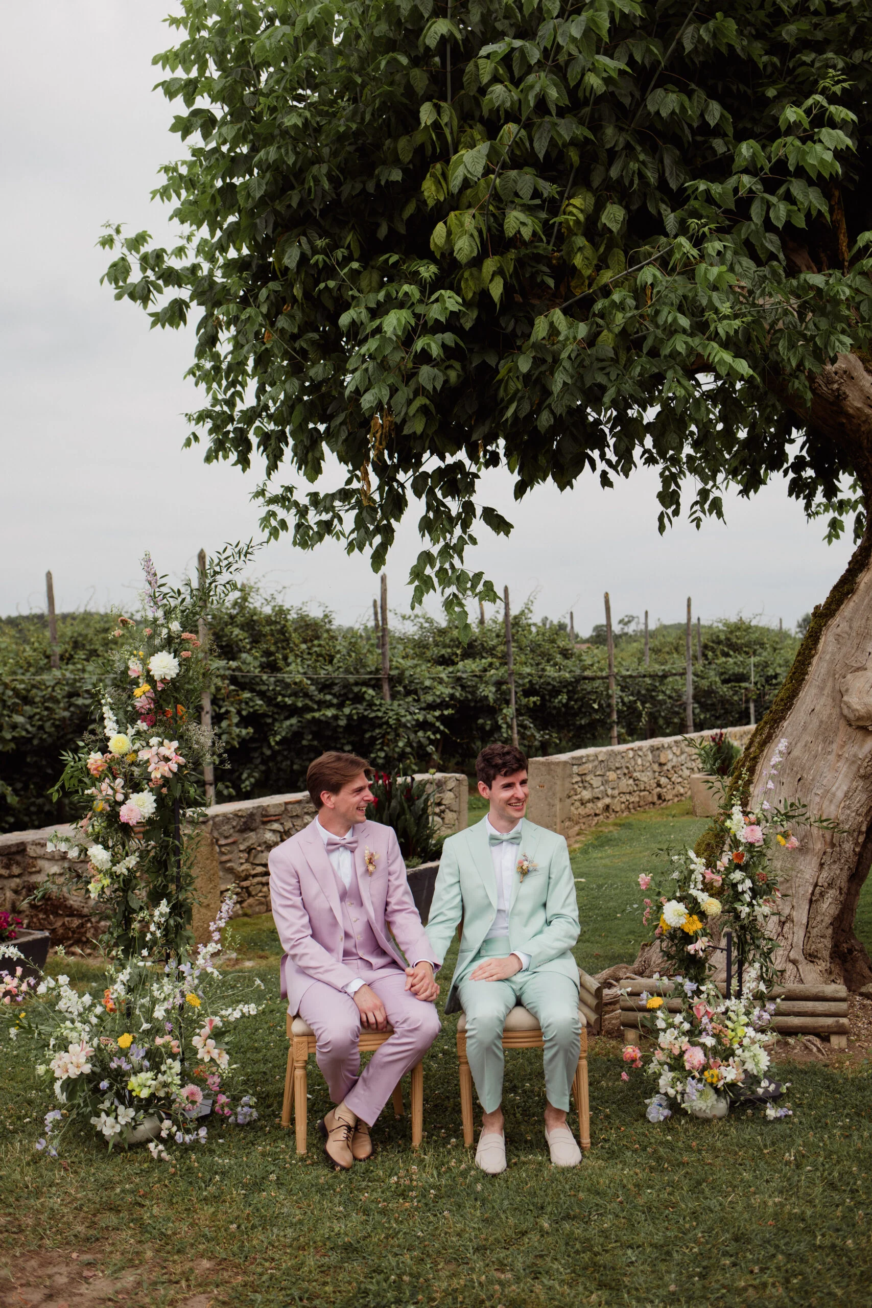 Deux hommes en costumes pastel rose et vert menthe assis côte à côte sous un arbre, entourés de fleurs champêtres.