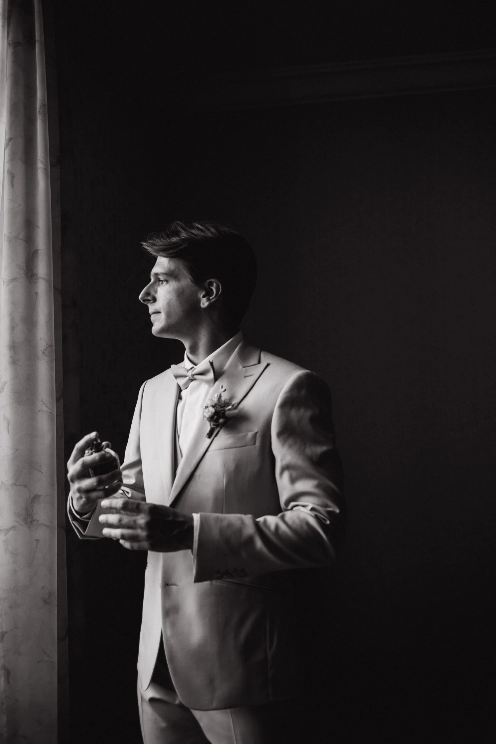Portrait en noir et blanc d'un marié en costume clair, se parfumant près d'une fenêtre dans une ambiance intimiste.