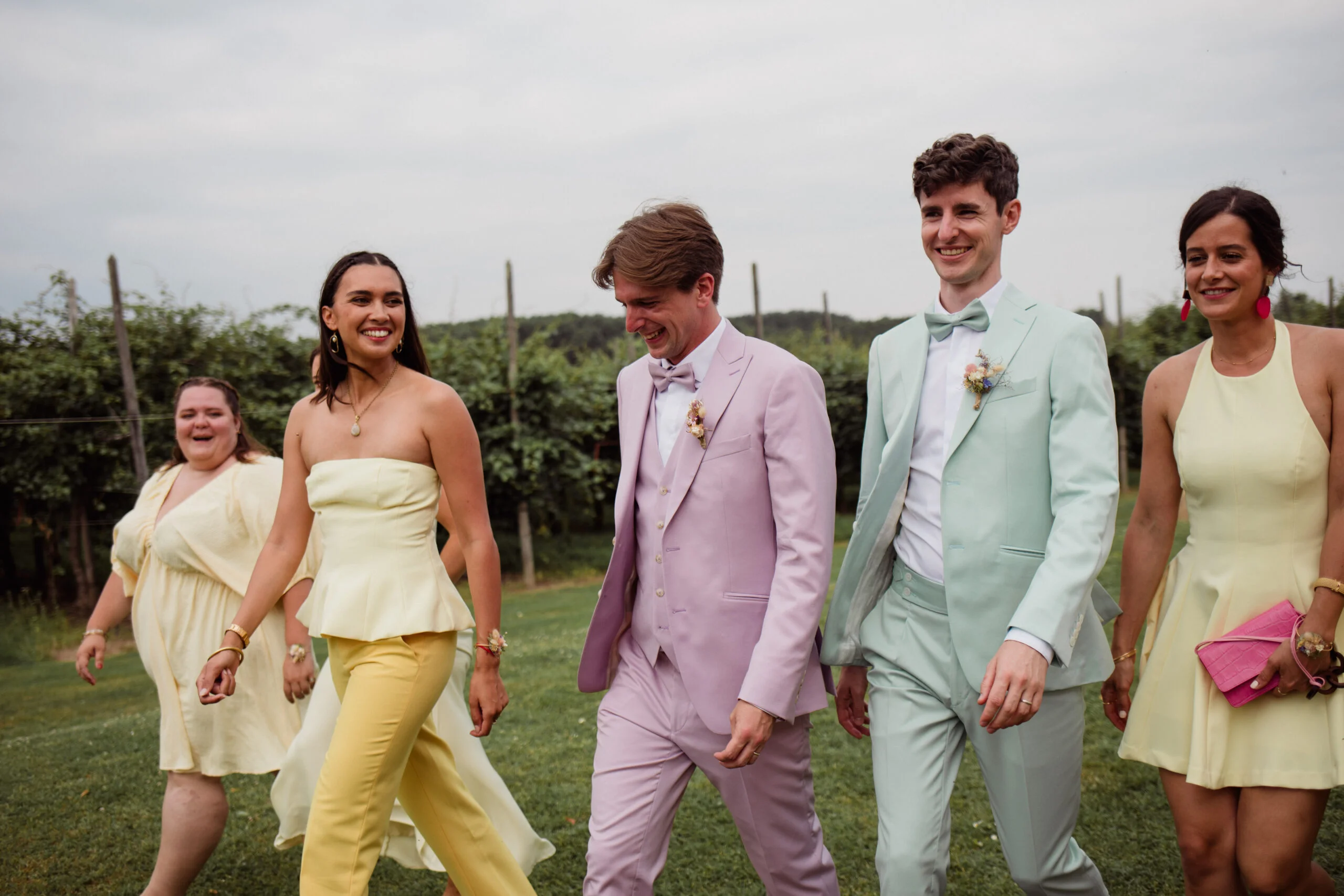 Quatre personnes souriantes marchent en extérieur, vêtues de costumes et robes aux tons pastel jaune, mauve et vert menthe.