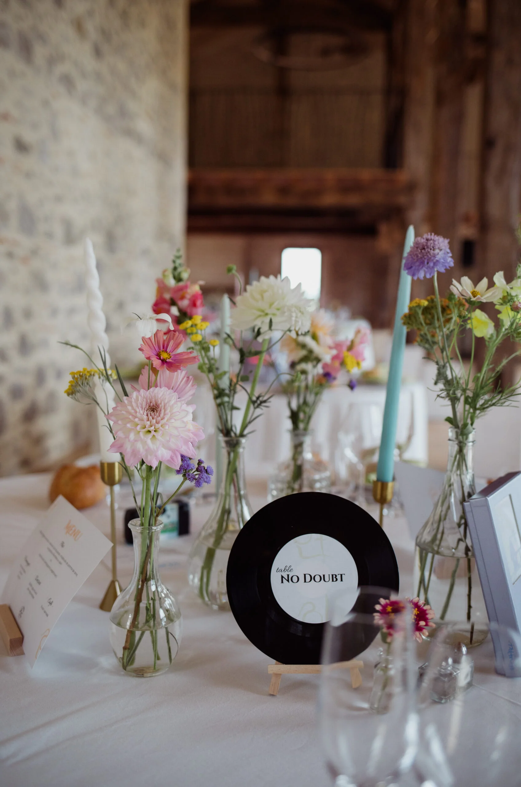 Décoration de table pastel avec fleurs champêtres, bougies et un vinyle servant de marque-place dans une grange.
