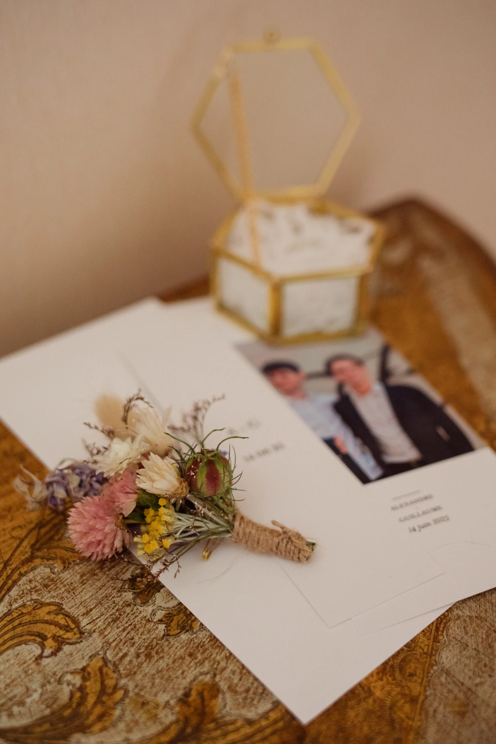 Boutonnière aux tons pastel posée sur un faire-part et une boîte à alliances dorée sur un meuble en bois rustique.
