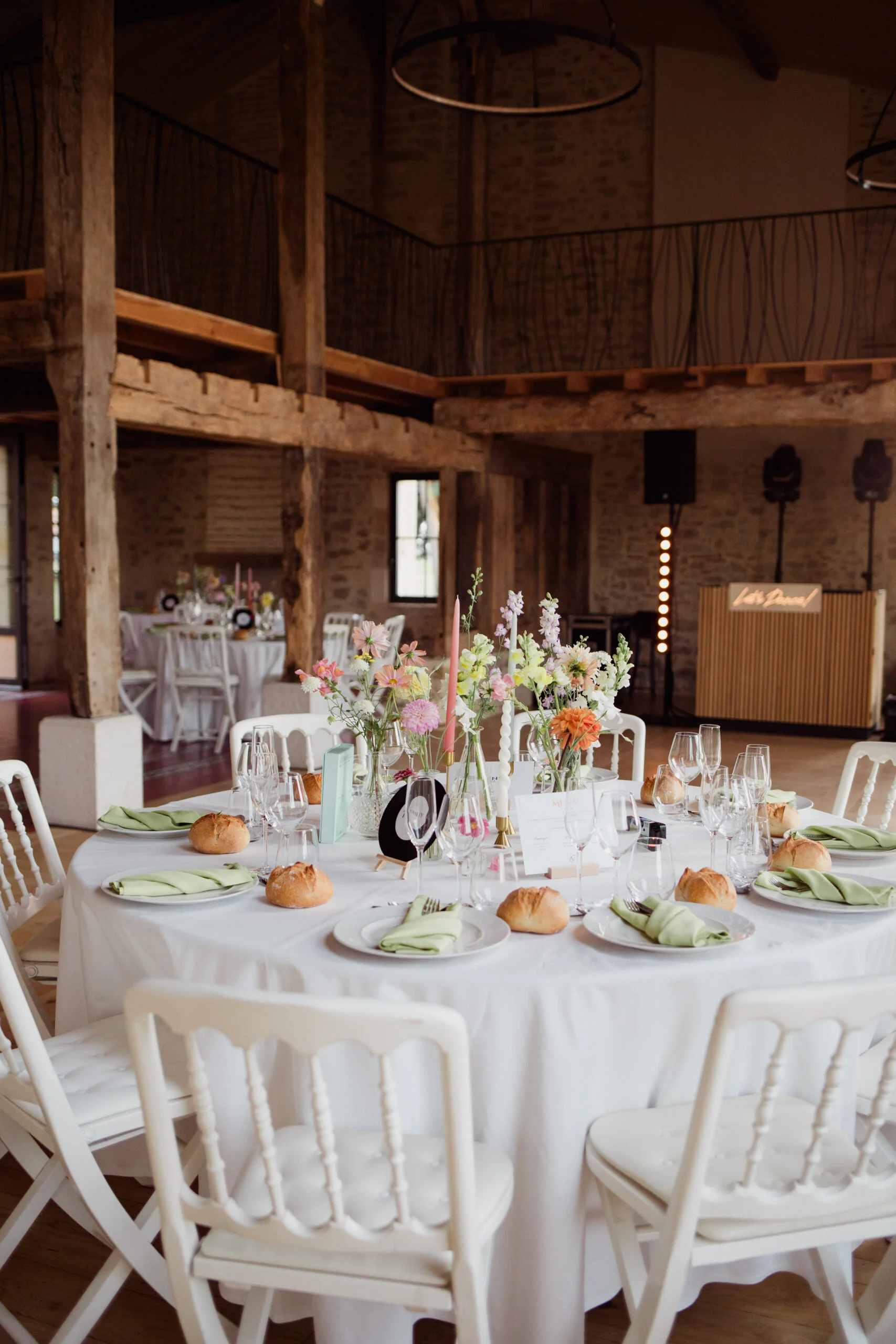Table ronde dressée avec nappage blanc, fleurs pastel et chaises blanches dans une grange rustique et lumineuse.