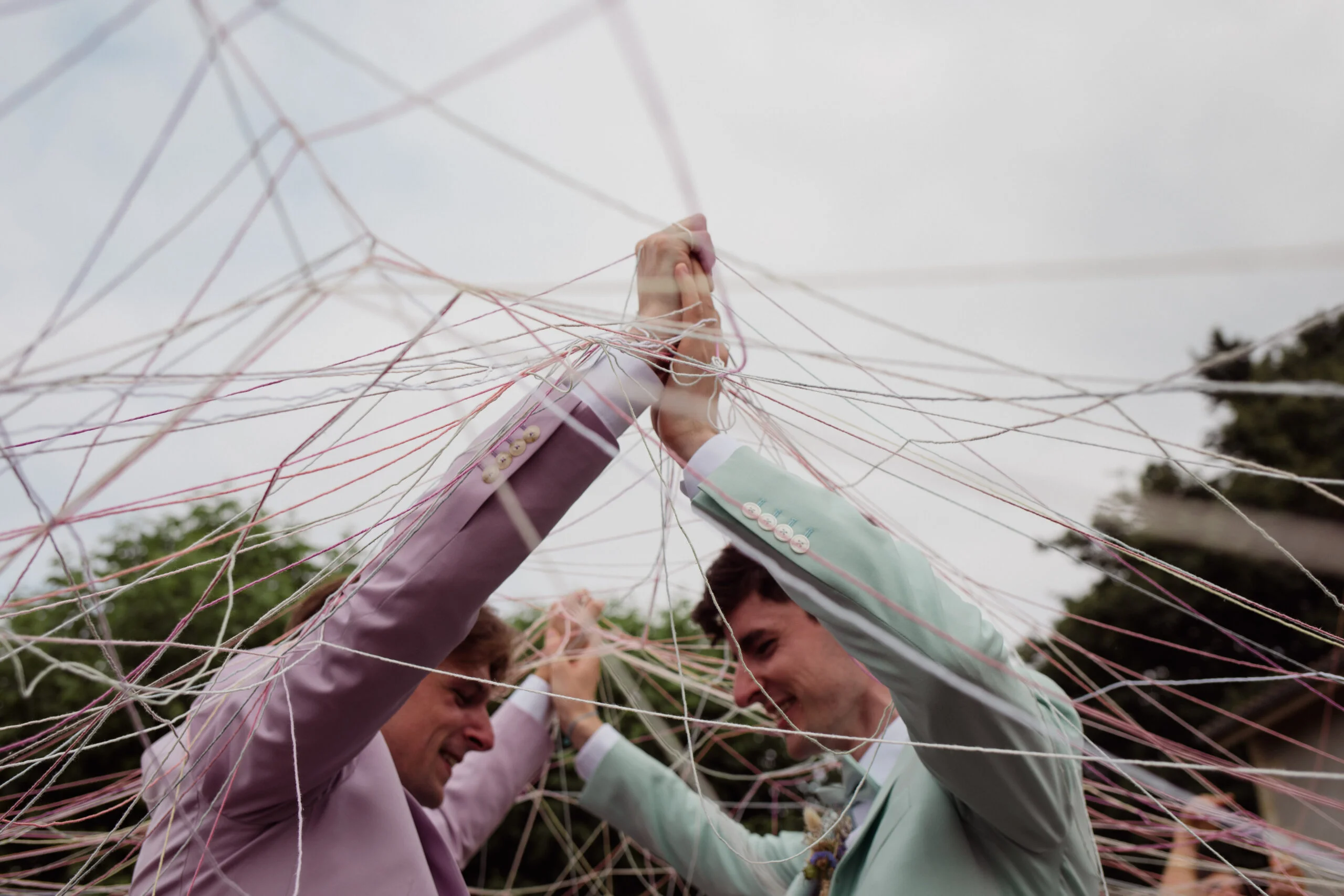 Deux hommes en costumes pastel se tiennent la main sous un réseau poétique de fils blancs et roses.