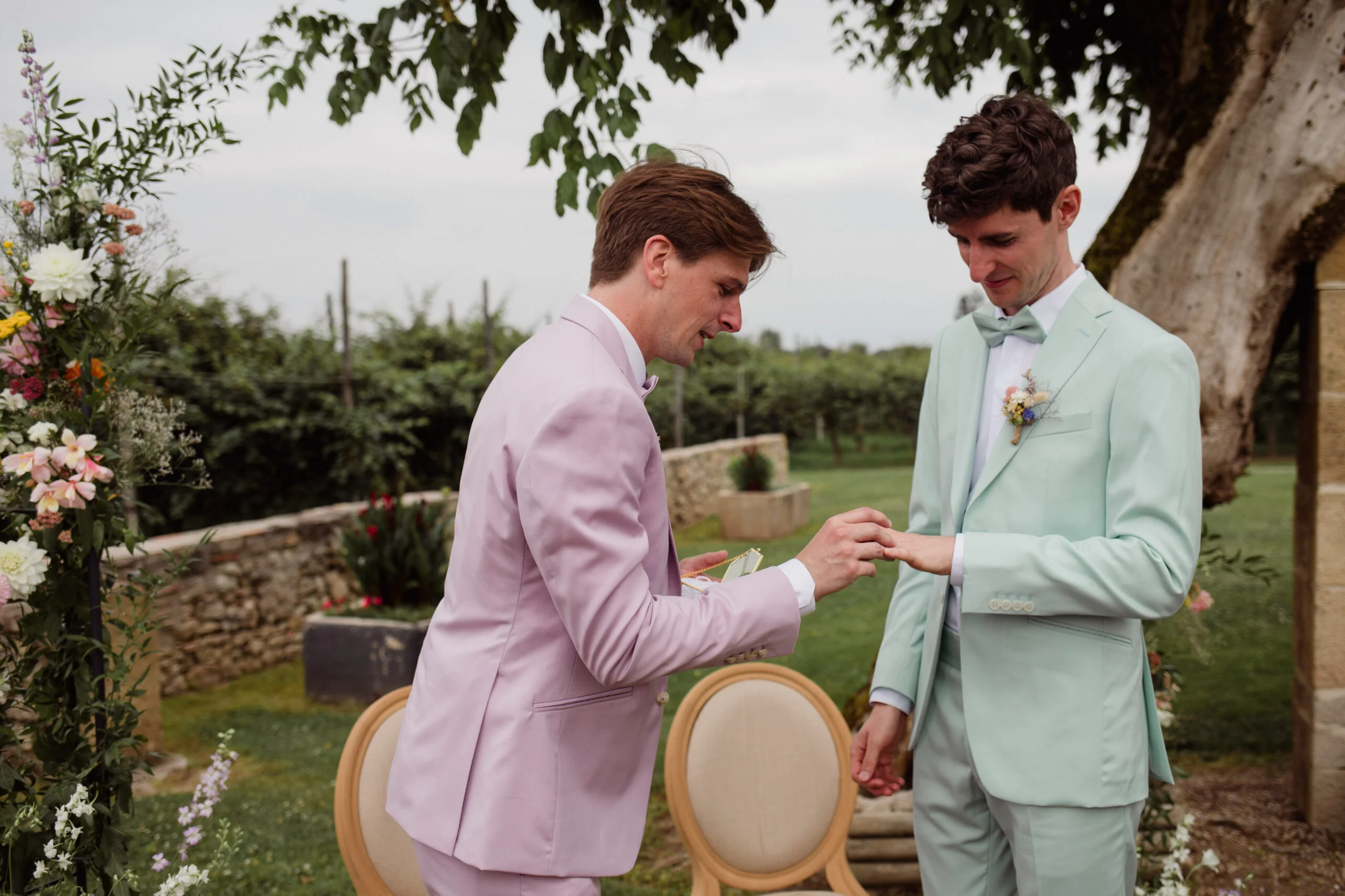 Deux hommes en costumes pastel, rose et vert menthe, s'échangent leurs alliances lors d'une cérémonie en plein air.