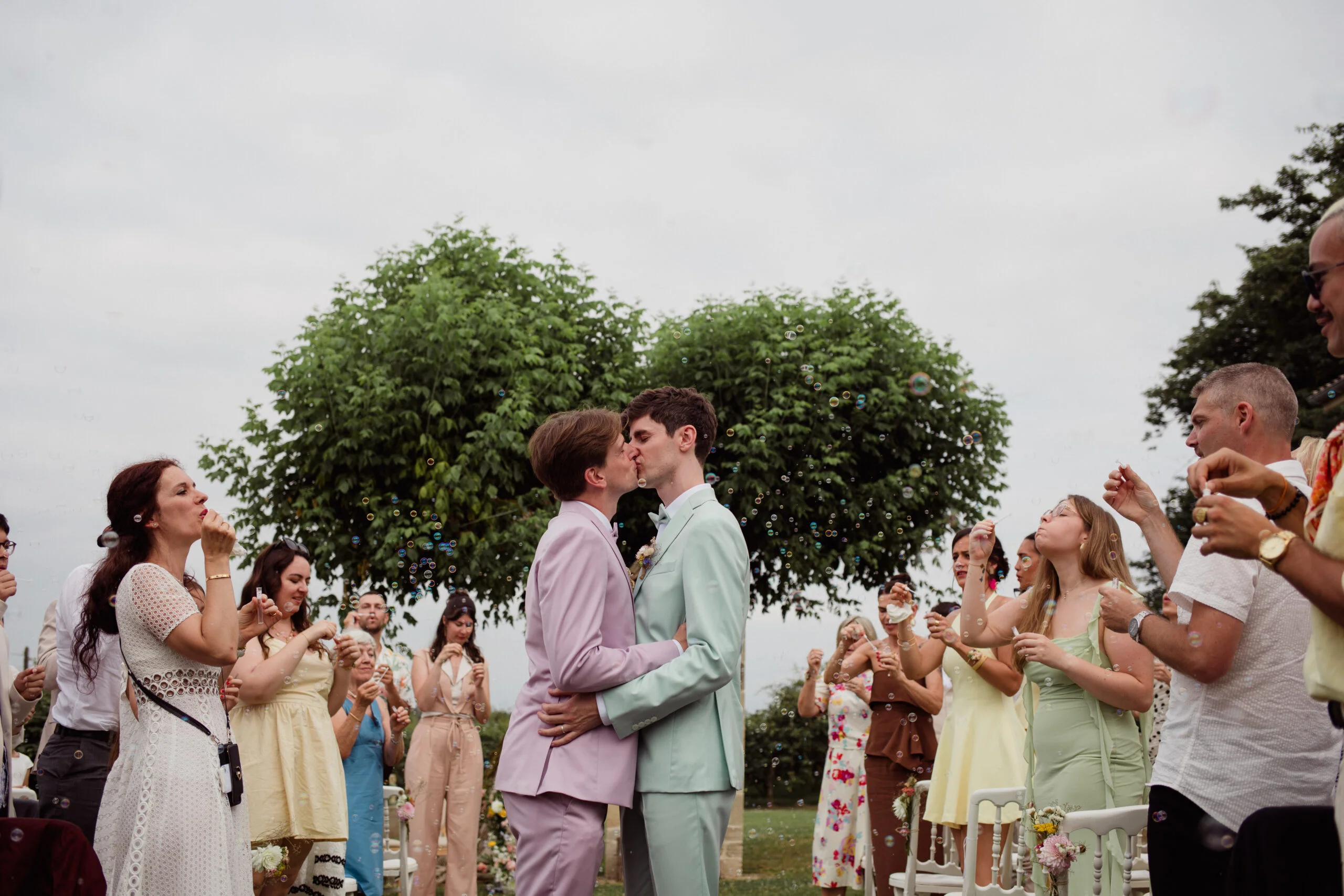 Deux hommes s'embrassent entourés d'invités et de bulles, vêtus de costumes pastel mauve et bleu clair en extérieur.