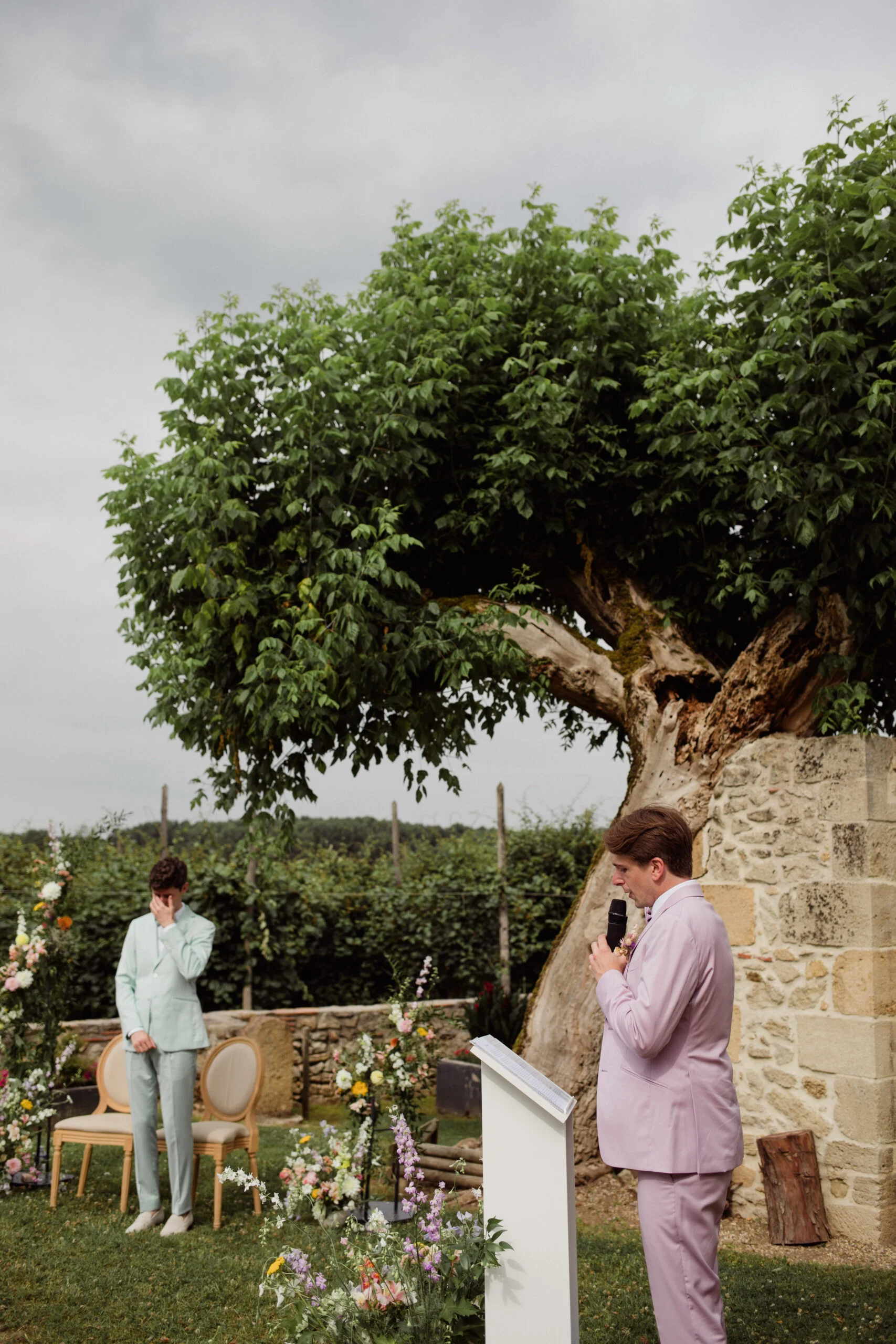 Deux hommes en costumes pastel s'adressent l'un à l'autre lors d'une cérémonie romantique sous un grand arbre.
