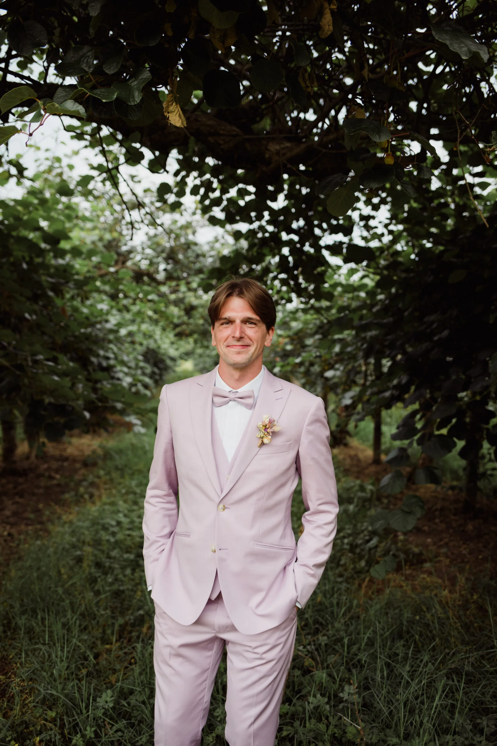 Un homme souriant en costume rose pastel avec nœud papillon, posant dans un jardin verdoyant et ombragé.