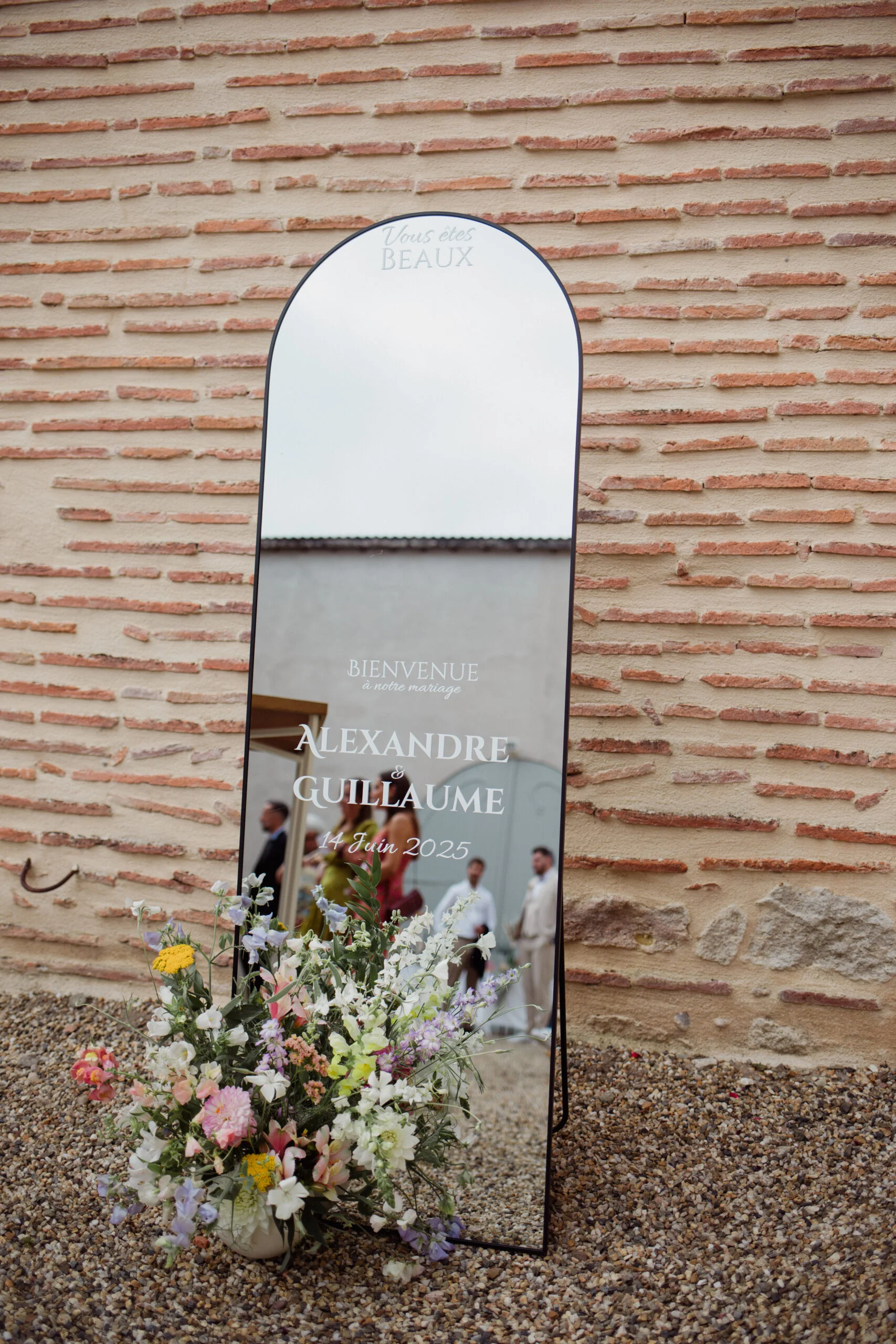 Miroir de bienvenue personnalisé avec fleurs pastel devant un mur de briques pour le mariage d'Alexandre et Guillaume.