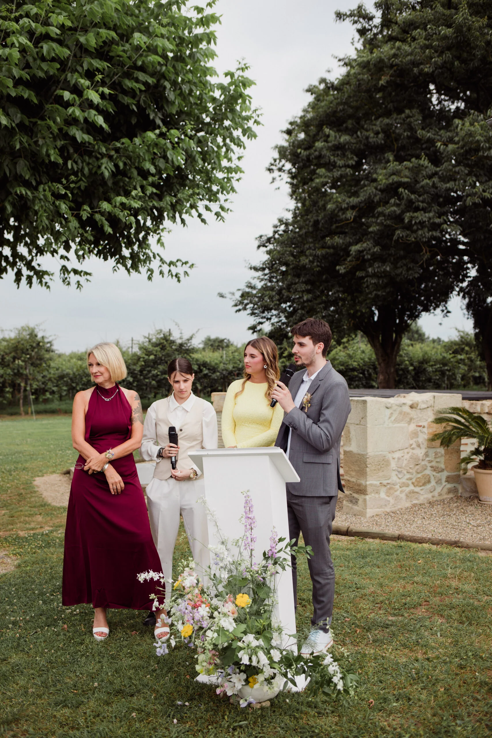 Trois personnes prononcent des discours lors d'une cérémonie en extérieur, entourées de fleurs et de verdure.