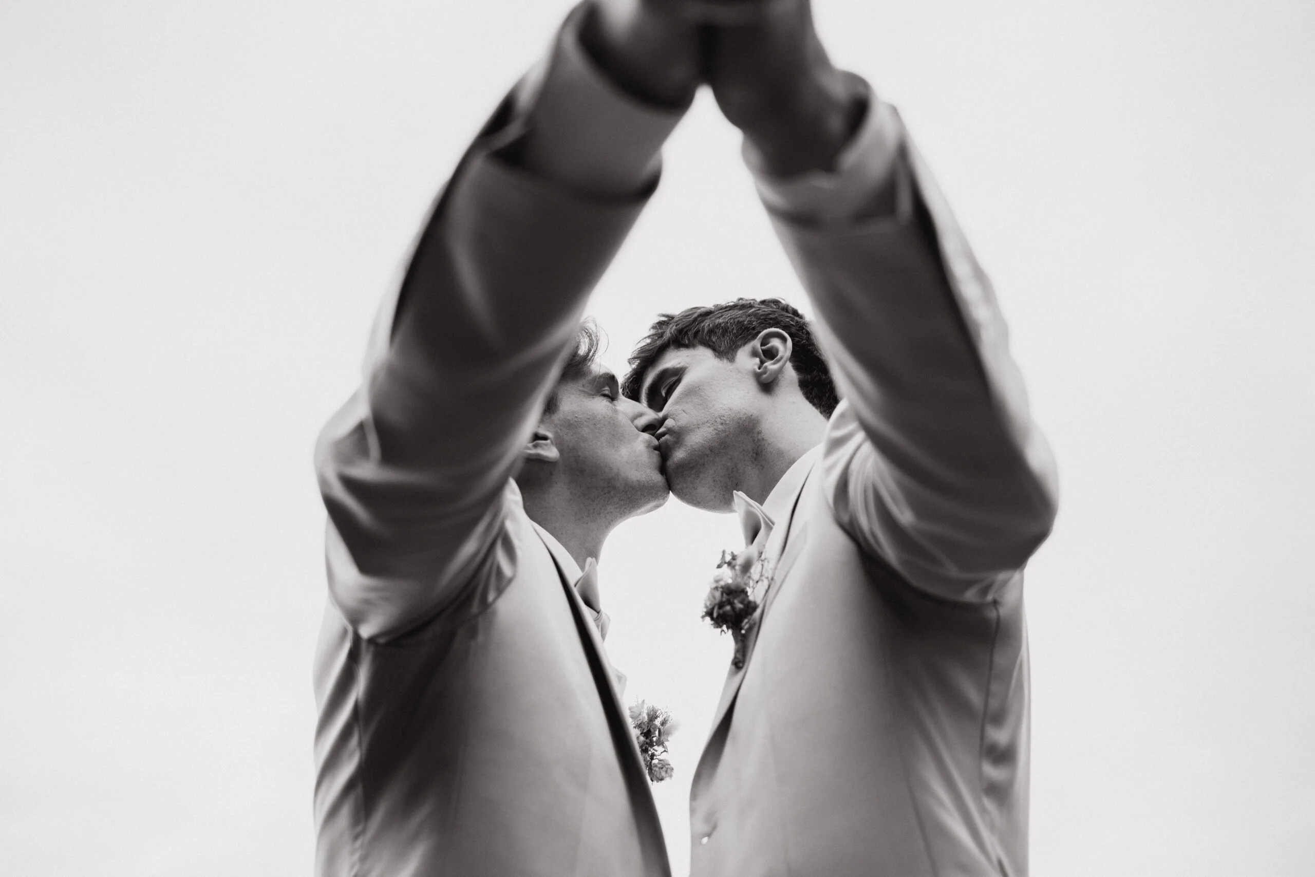 Deux hommes en costumes s'embrassent sous une arche formée par leurs bras, dans un cliché poétique en noir et blanc.