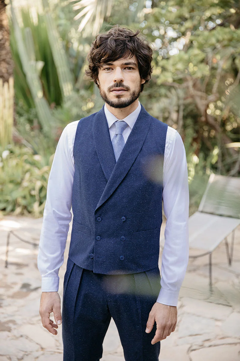 Un homme élégant dans un veston bleu avec boutons noirs, chemise blanche et cravate, se tient dans un jardin luxuriant.