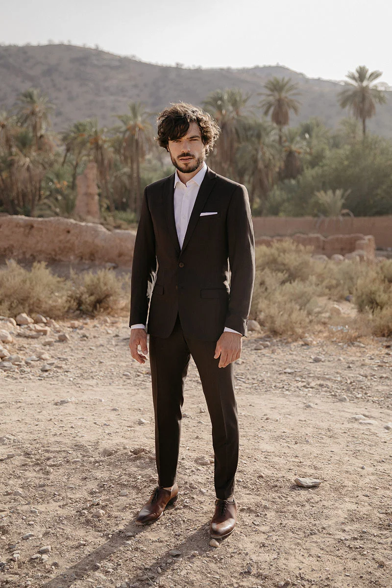 Un homme élégant dans un costume noir classique se tient fièrement dans un décor rustique, incarnant la mode masculine