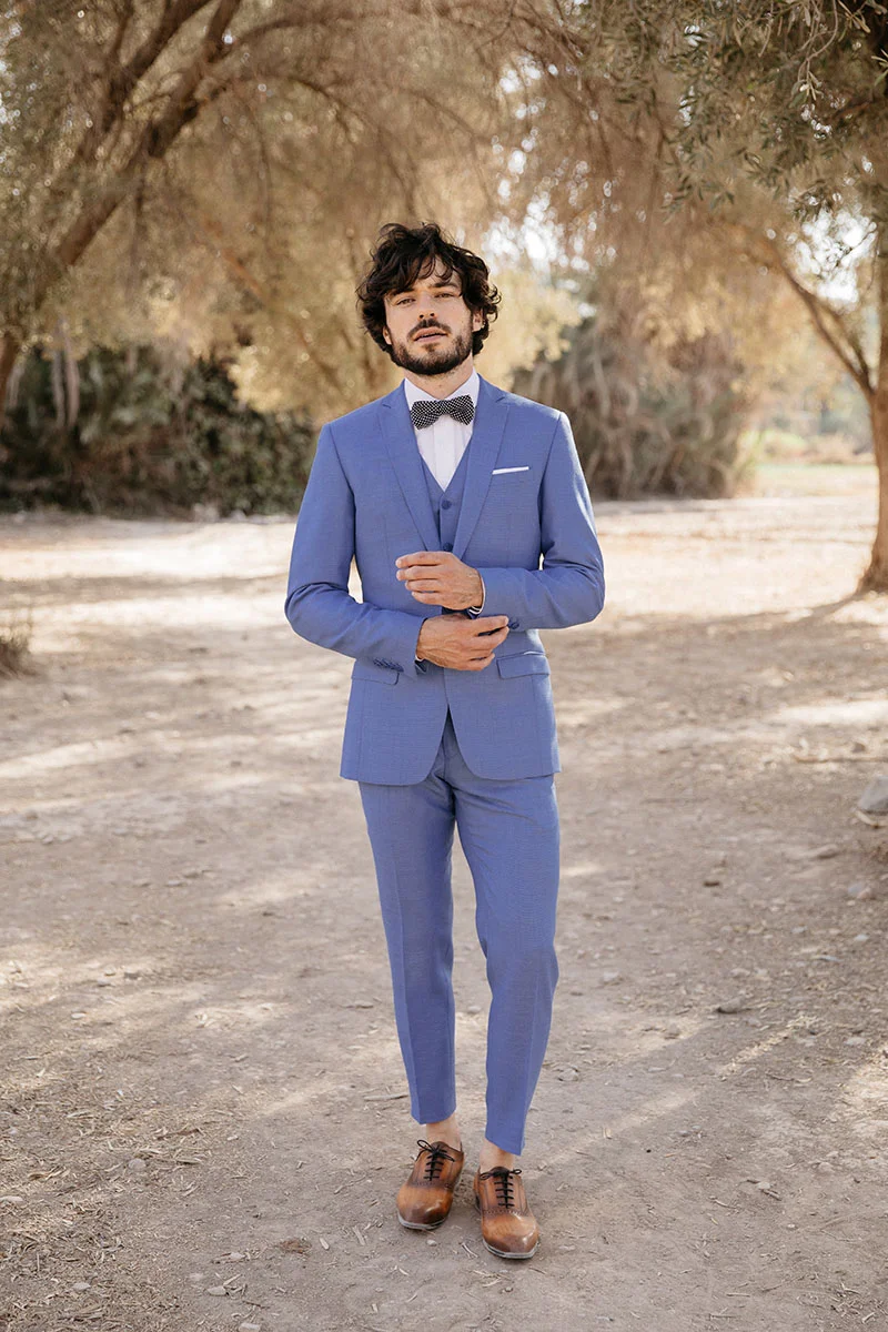 Un homme élégant dans un costume bleu clair et une cravate noire avec des motifs blancs, posant dans un parc paisible.
