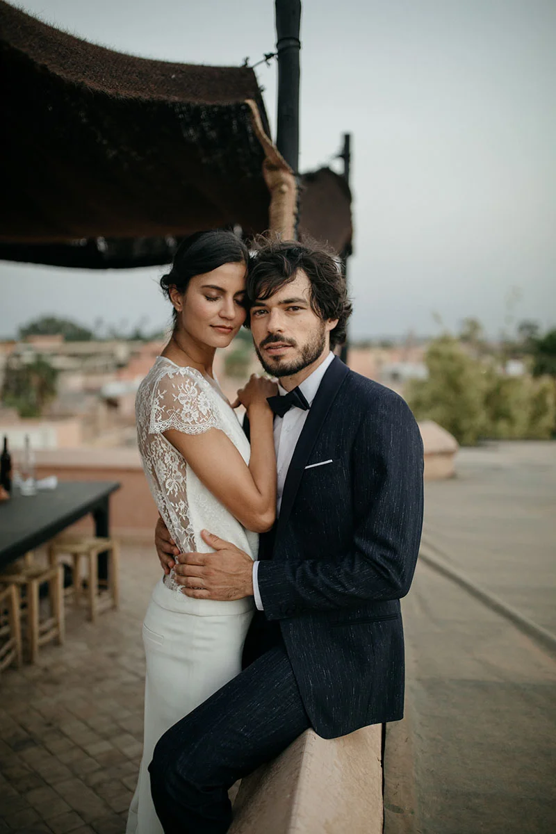 Un couple romantique dans des vêtements de mariage chic et sophistiqués, se tenant sur un balcon avec un arrière-plan paisible.