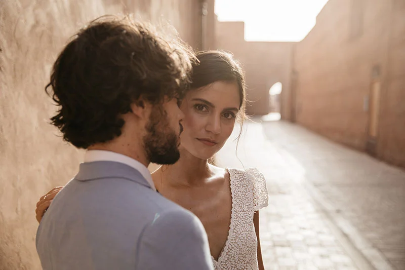 Un couple romantique dans une rue ensoleillée, l'homme en costume bleu, la femme en robe de mariage blanche, incarnant