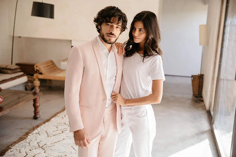 Un couple chic dans des costumes rose et blanc dans un salon moderne, incarnant l'élégance et l'originalité pour un mariage.