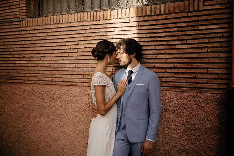 Un couple romantique se détient contre un mur en briques, l'homme en costume bleu et l'homme en robe blanche, incarnant