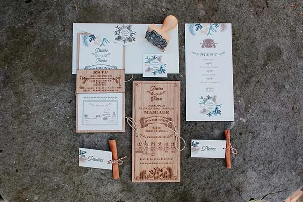 Papeterie de mariage champêtre en bois et papier blanc avec motifs floraux bleus, posée sur un fond minéral gris.