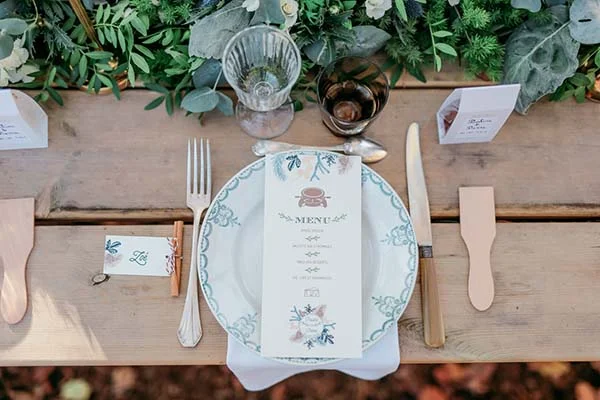 Dressage de table champêtre avec menu, couverts dorés et feuillage vert sur une table en bois clair.