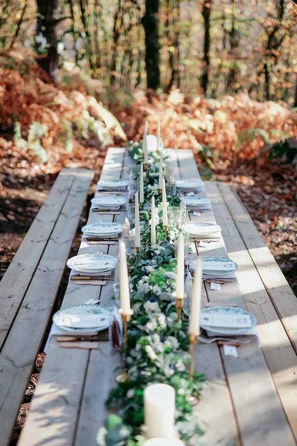 Table champêtre en bois décorée de bougies et de feuillage vert, dressée en plein air dans une forêt automnale.