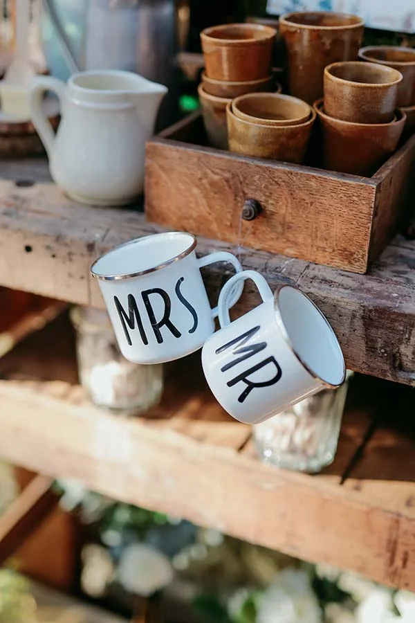 Deux tasses blanches marquées Mr et Mrs suspendues à un étalage en bois rustique dans une ambiance champêtre.