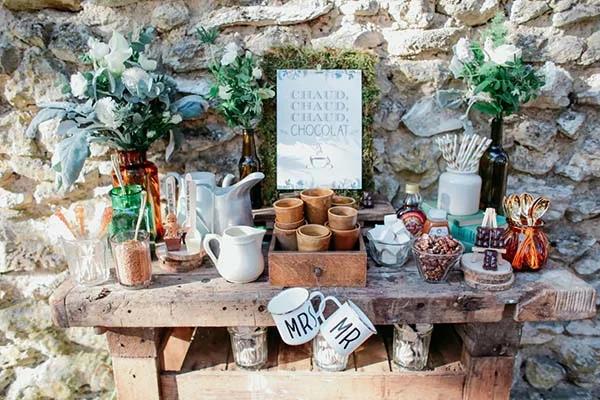 Bar à chocolat chaud champêtre avec tasses personnalisées Mr & Mrs, fleurs blanches et décor rustique en bois.