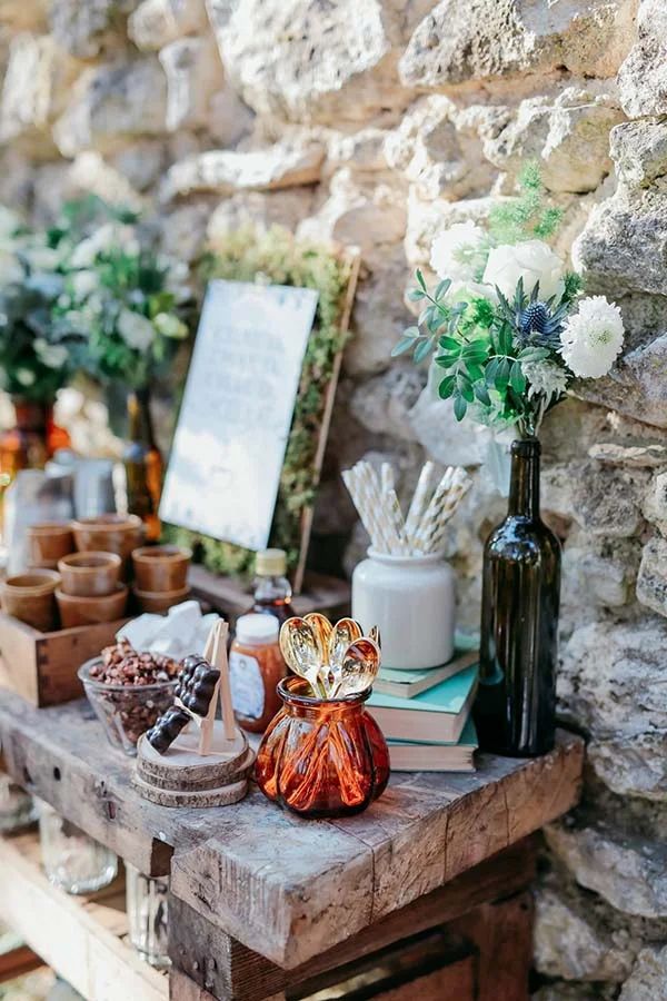 Buffet champêtre avec fleurs blanches, bouteilles en verre et accessoires naturels devant un mur de pierres anciennes.