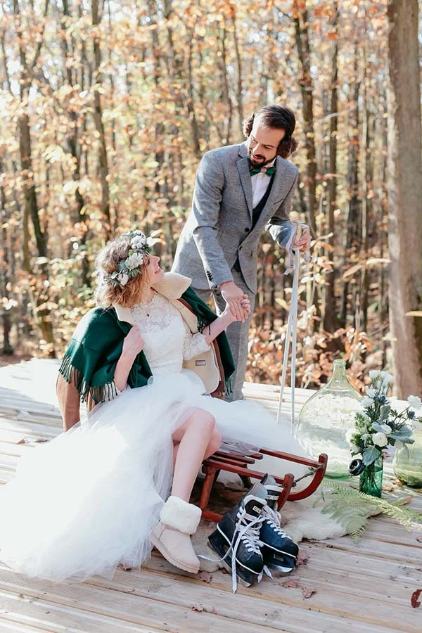 Couple de mariés en tenue élégante et hivernale posant avec complicité sur un traîneau en bois dans une forêt d'automne.