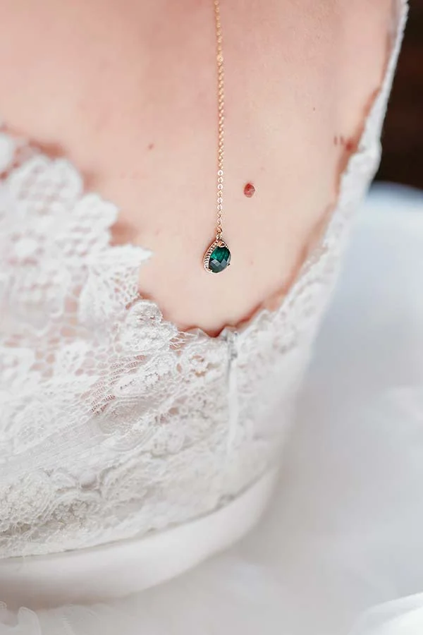 Détail du dos d'une robe de mariée en dentelle blanche avec un fin collier doré et un pendentif vert émeraude.