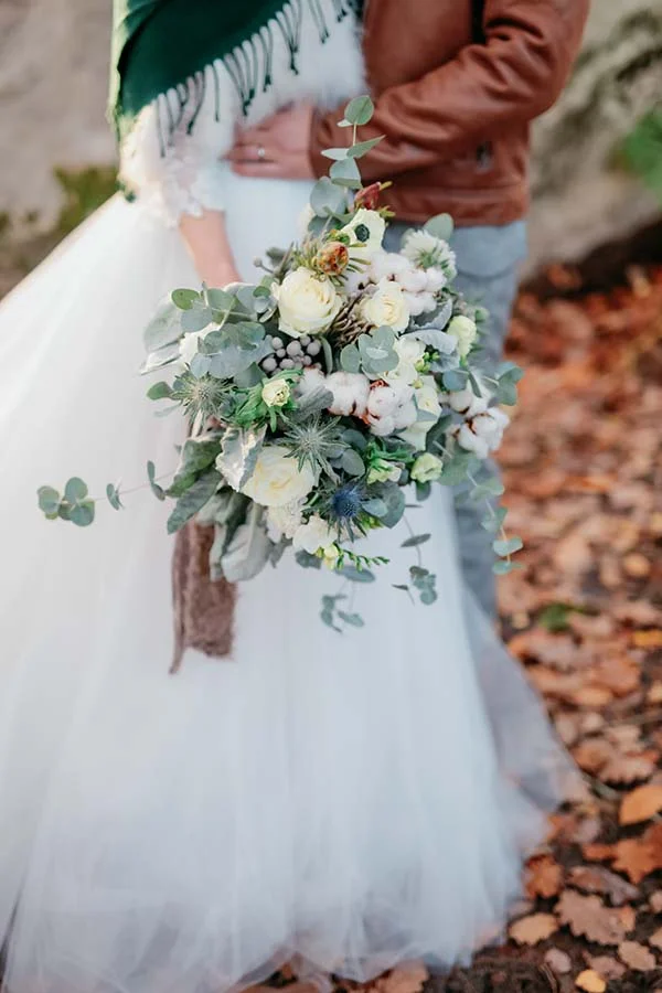 Gros plan d'un bouquet champêtre blanc et eucalyptus tenu par des mariés dans un décor automnal.