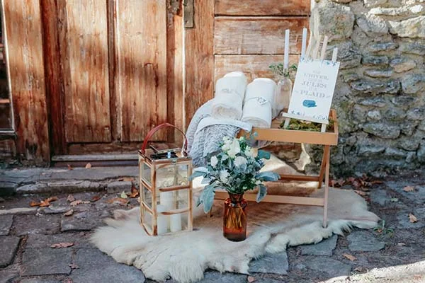 Coin cosy champêtre avec plaids blancs, lanterne et fleurs sur une peau de mouton devant un mur en bois et pierre.