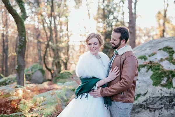 Couple marié en tenue hivernale, elle en robe blanche et étole, lui en veste cuir, dans une forêt automnale lumineuse.