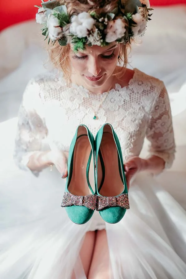 Mariée en robe de dentelle et couronne de fleurs tenant une paire d'escarpins verts à paillettes.
