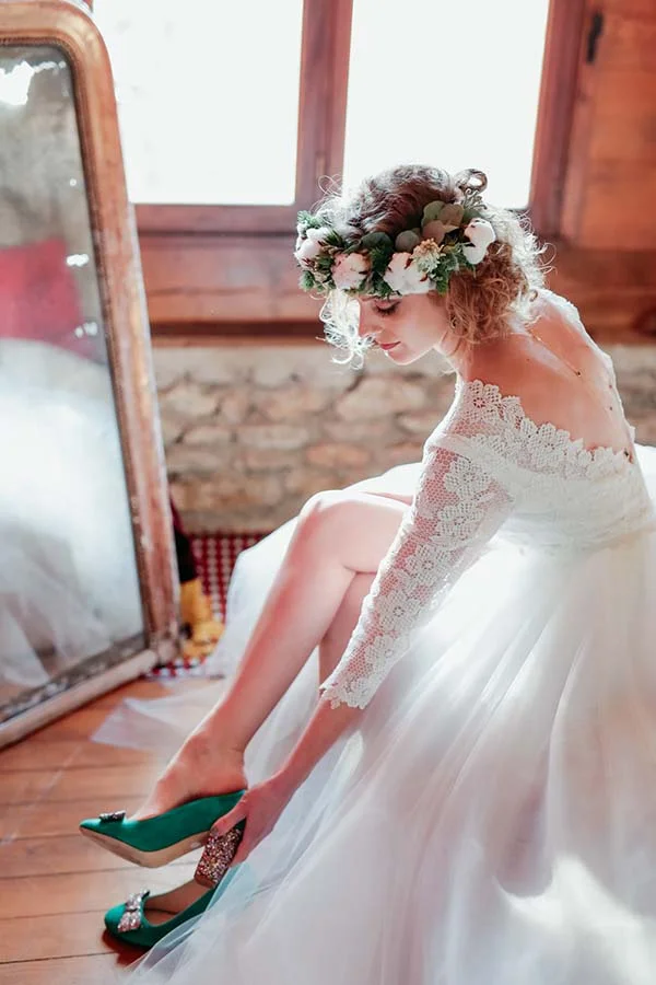 Mariée en robe blanche et couronne de fleurs, chaussant des escarpins verts dans un intérieur rustique et lumineux.