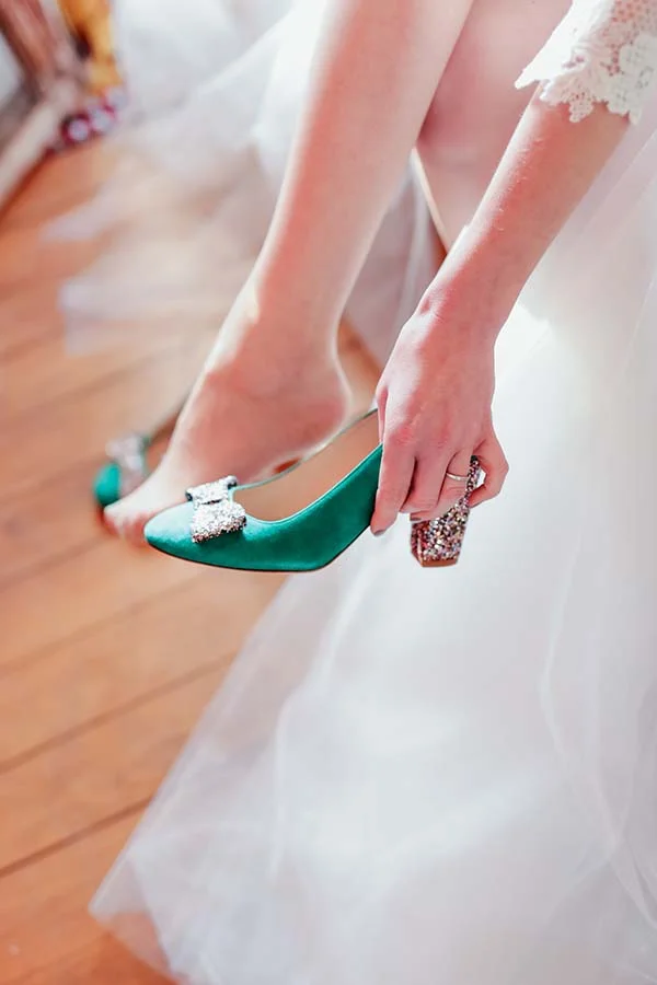 Une mariée en robe blanche enfile des escarpins verts ornés de bijoux pour une touche d'originalité.