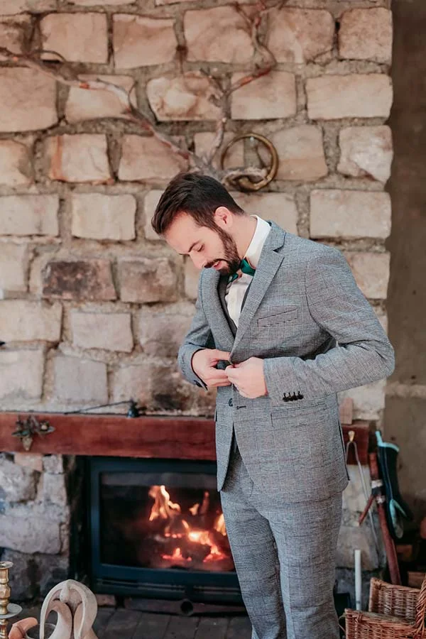 Marié en costume gris ajusté boutonnant sa veste devant une cheminée rustique en pierre.