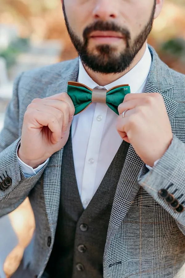 Gros plan d'un marié ajustant son nœud papillon vert et beige, portant un costume gris à carreaux et un gilet sombre.