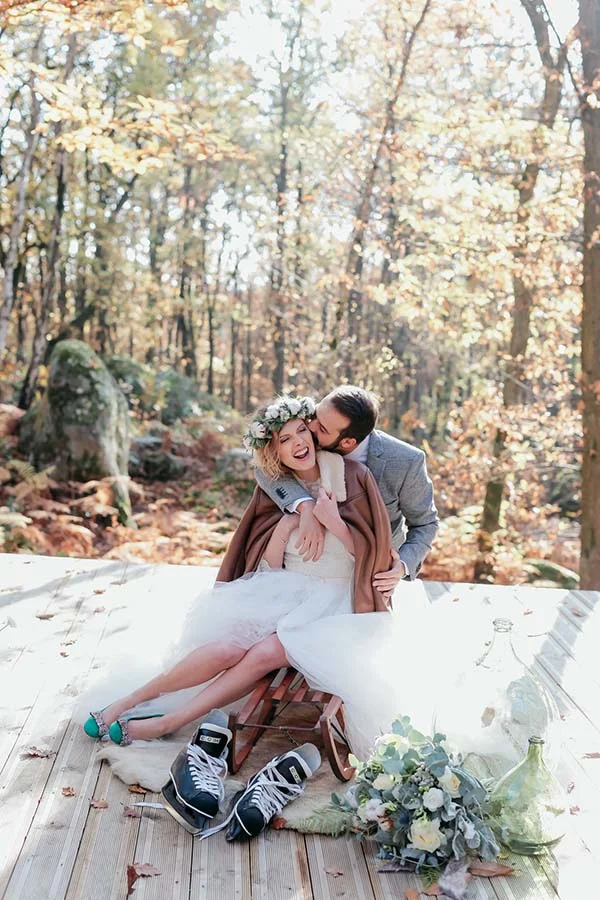 Couple marié en tenue champêtre s'embrassant tendrement sur une terrasse en bois dans une forêt aux couleurs d'automne.