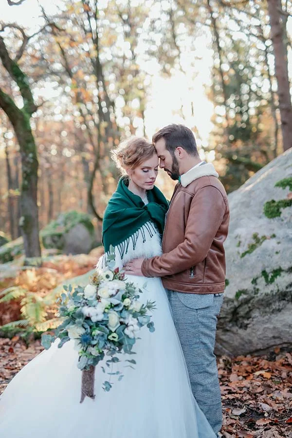 Couple marié s'enlaçant tendrement en forêt d'automne, elle en robe blanche et châle vert, lui en veste en cuir marron.