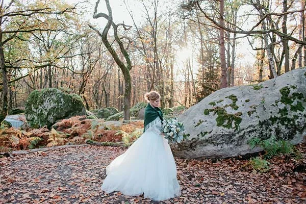 Mariée en robe blanche et étole verte tenant un bouquet, posant dans une forêt automnale aux tons chauds.