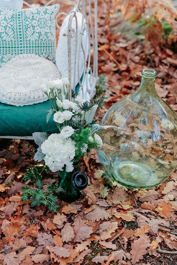 Décoration champêtre avec fleurs blanches, dame-jeanne en verre et coussins sur un tapis de feuilles d'automne.