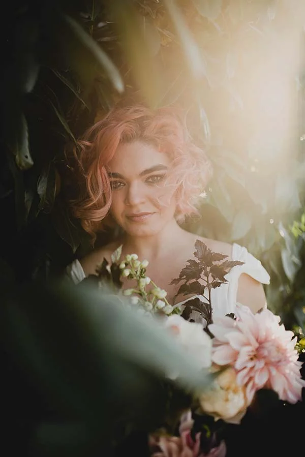 Une jeune femme avec des cheveux roses tendres se tient dans un décor verdoyant, entourée de fleurs blanches et roses,