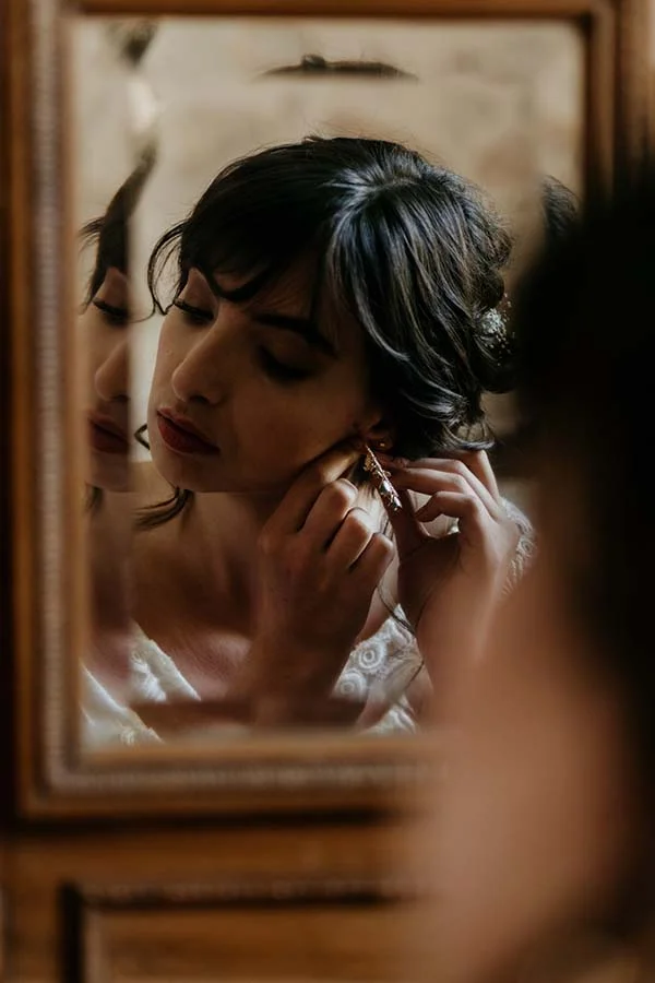 Élégante mariée en robe blanche, ajustant ses bijoux dans un miroir vintage, préparant sa journée de mariage nature et