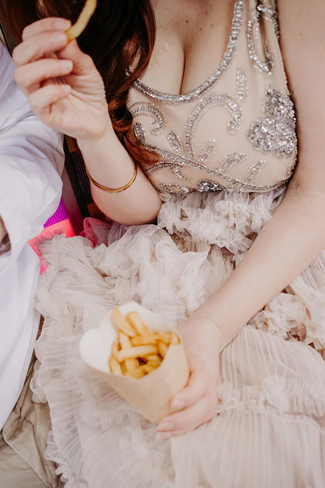 Une femme en robe époustouflante de mariage déguste des frites dans un décor romantique et luxueux.