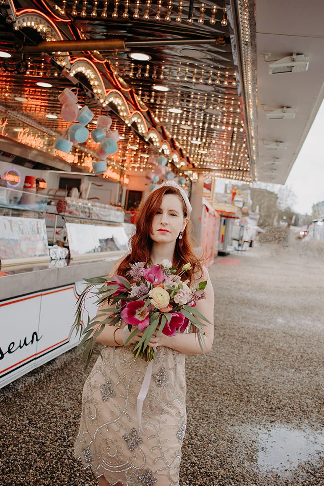 Une jeune femme dans un robe époustouflante tient un bouquet de fleurs vibrantes contre un décor de cirque lumineux.