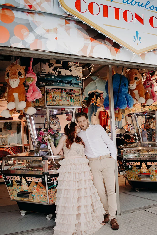 Un couple charmant dans un décor de foire, avec un arrière-plan coloré et joyeux, capturant l'essence d'une expérience