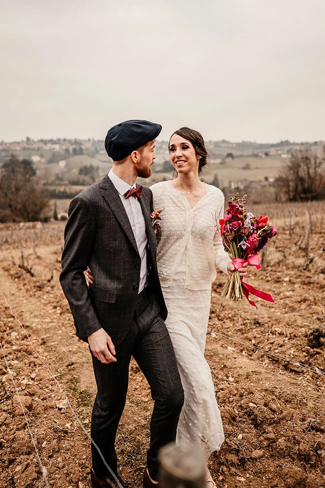 Un couple charmant dans des vêtements vintage se promène dans un champ, le mari tenant un bouquet magenta.