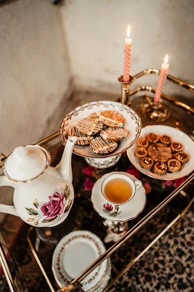 Un déjeuner romantique avec une théière à motifs fleuris, des biscuits et des tarts, entouré de candelles douces et de