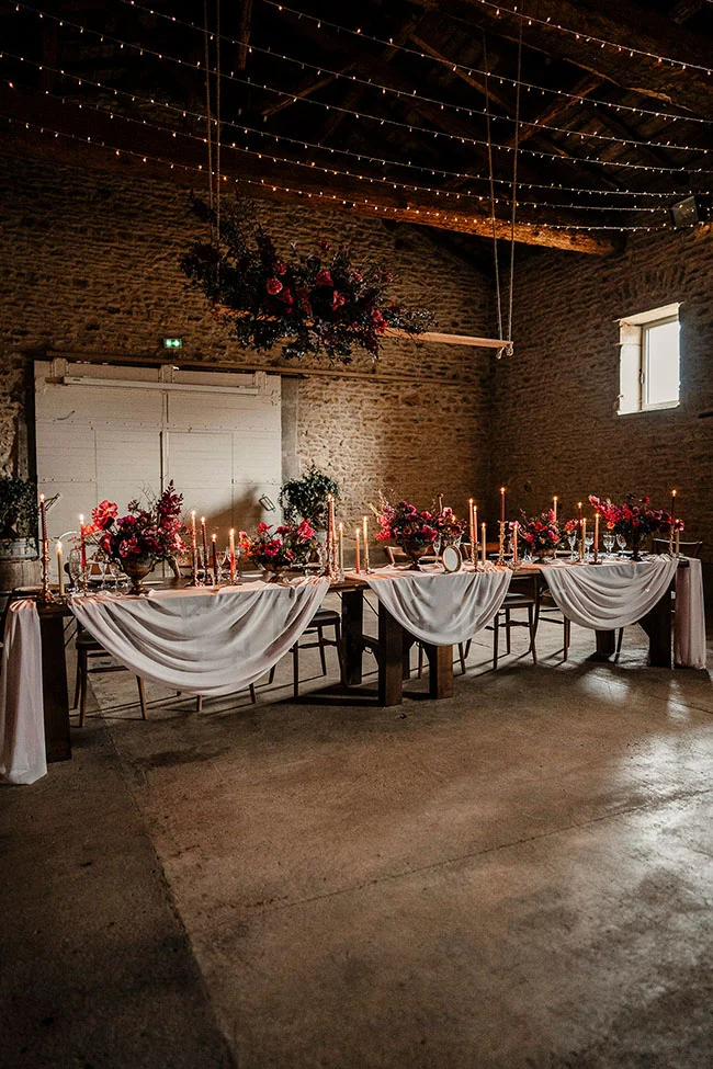 Salle rustique éclairée d'une lumière chaude, avec une table de banquet ornée de cierpes blanches et de fleurs magenta,