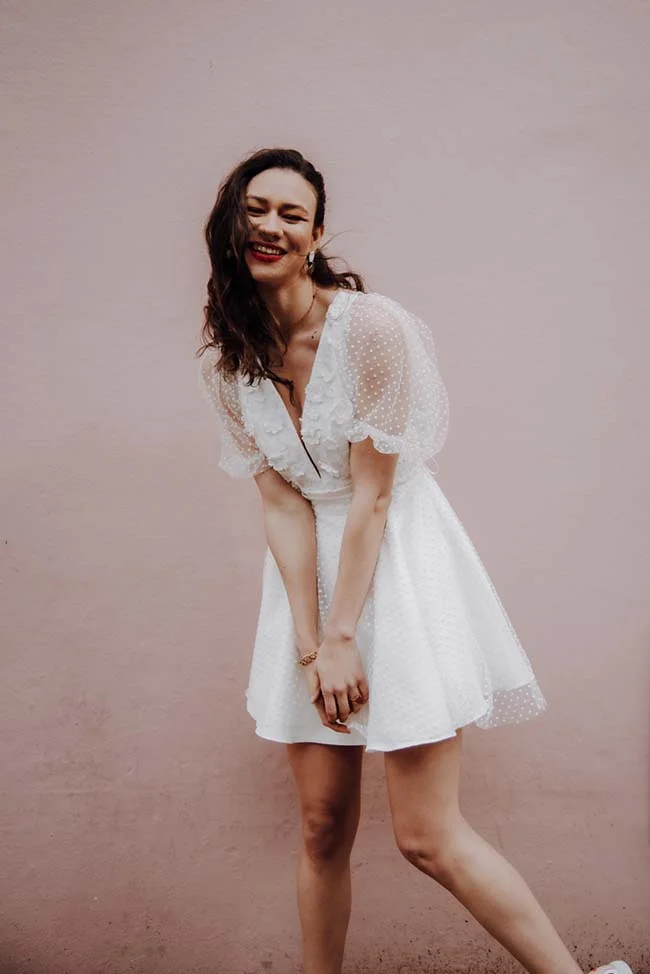 Une jeune femme dans un joli robe blanche se contorte joyeusement contre un mur rose, incarnant l'essence d'une journée